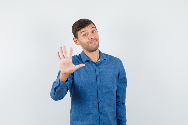 Giovane che fa il segnale di stop educatamente in camicia blu e sembra allegro, vista frontale.