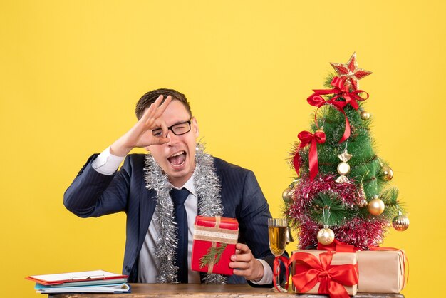 giovane che fa segno giusto davanti all'occhio seduto al tavolo vicino albero di Natale e regali su giallo