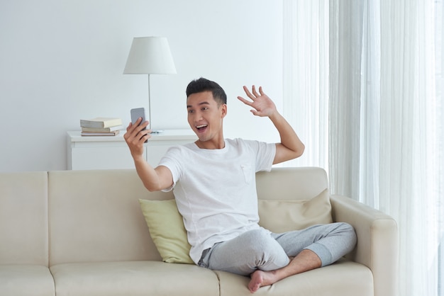 Giovane che fa una videochiamata che si siede sul sofà nel suo salone e che dà un gesto dell'onda di saluto