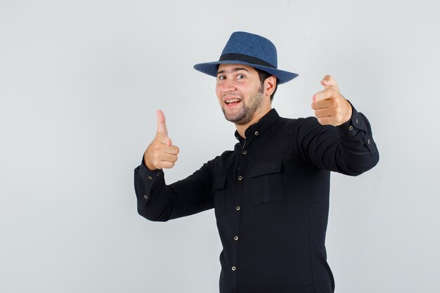 Giovane che indica alla macchina fotografica con il gesto della pistola in camicia nera, cappello e che sembra allegro.