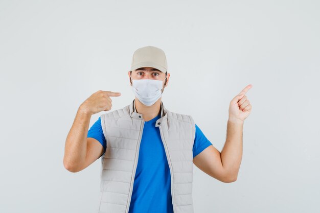 Giovane che indica la sua maschera e su in t-shirt, giacca, berretto e sembra serio, vista frontale.