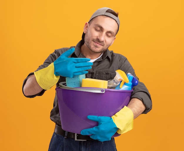 Giovane che indossa abiti casual e berretto in guanti di gomma che tiene secchio con strumenti di pulizia inalando piacevole aroma fresco in piedi sopra la parete arancione