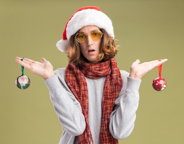 Giovane che indossa il cappello di Babbo Natale e occhiali gialli con una sciarpa calda intorno al collo che tiene le palle di Natale confuse in piedi sul muro verde