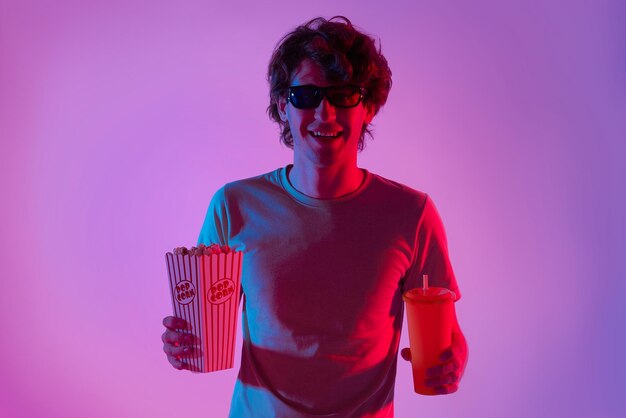 Giovane che indossa occhiali 3D che si diverte a mangiare popcorn e guardare film su sfondo di luci al neon