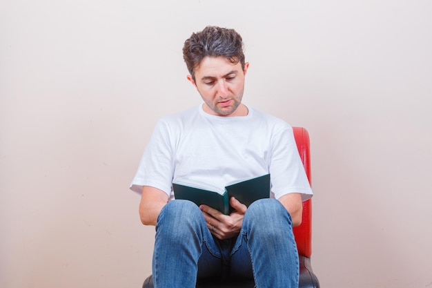 Giovane che legge un libro mentre è seduto su una sedia in t-shirt, jeans