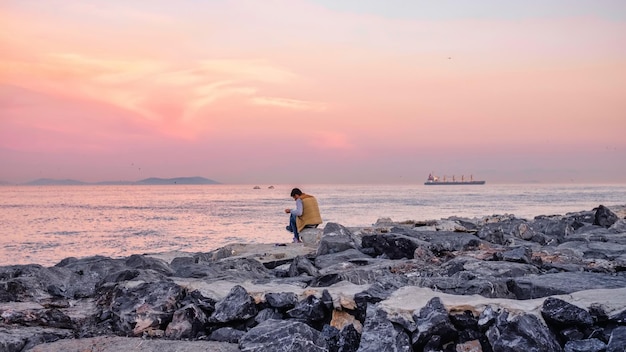 Giovane che medita e legge sulla riva di una spiaggia una barca che passa al tramonto