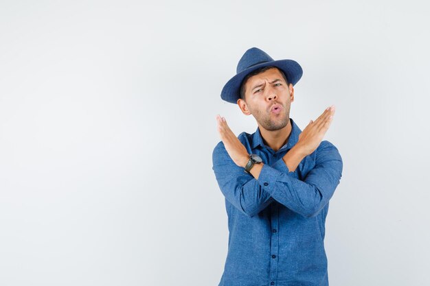 Giovane che mostra gesto di arresto in camicia blu, cappello, vista frontale.