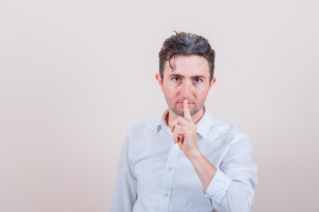 Giovane che mostra gesto di silenzio in camicia bianca e guarda attento