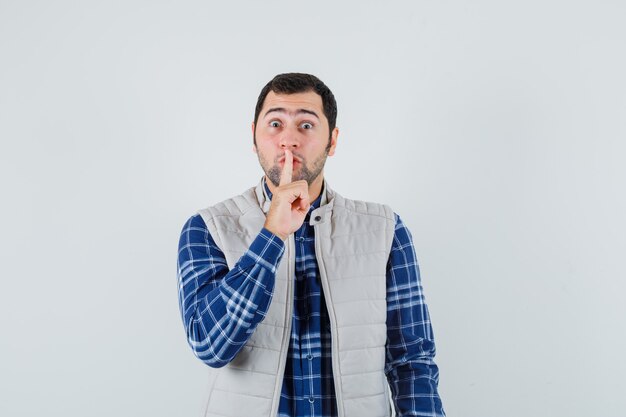 Giovane che mostra gesto di silenzio in camicia, giacca senza maniche e che sembra calmo. vista frontale.