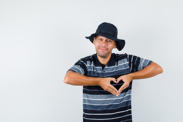 Giovane che mostra il gesto del cuore in maglietta, cappello e sembra allegro, vista frontale.