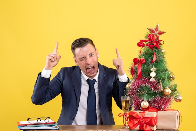 giovane che mostra la lingua che indica con il dito in su seduto al tavolo vicino all'albero di Natale e regali su giallo