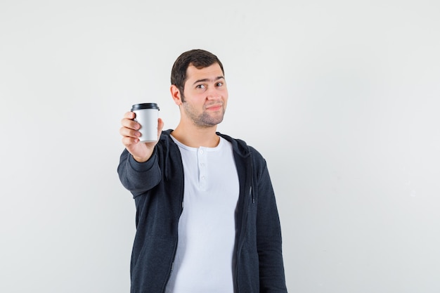 Giovane che mostra la tazza di caffè da asporto alla telecamera in t-shirt bianca e felpa con cappuccio nera con zip e sembra ottimista, vista frontale.