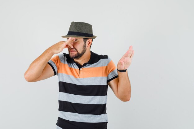 Giovane che pizzica il naso a causa del cattivo odore in t-shirt, cappello e sembra disgustato, vista frontale.