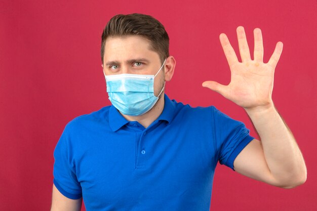 Giovane che porta la camicia di polo blu nella maschera protettiva medica che fa gesto di saluto con l'ondeggiamento aperto della mano che controlla parete rosa isolata