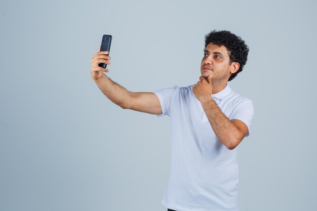 Giovane che posa mentre prende selfie in maglietta bianca e sembra carino. vista frontale.