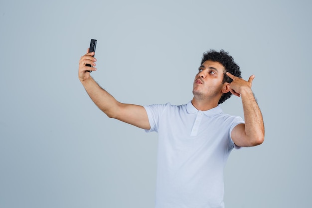 Giovane che posa mentre prende selfie sul telefono cellulare in maglietta bianca e sembra sicuro, vista frontale.