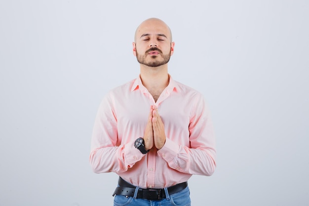 Giovane che prega in camicia rosa, jeans e guardando fiducioso, vista frontale.