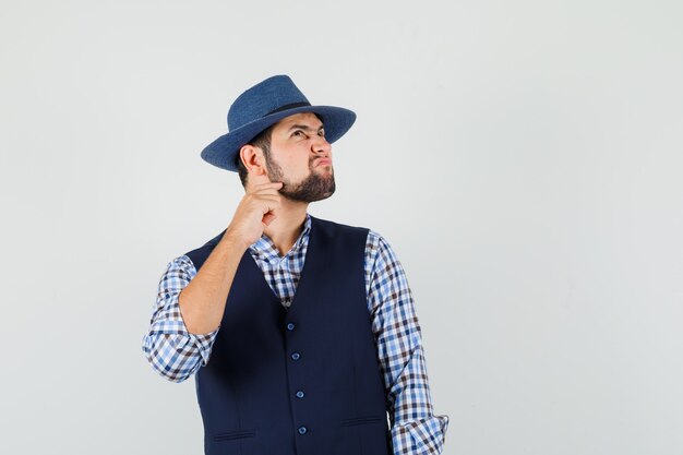 Giovane che si gratta la barba in camicia, gilet, cappello e che sembra pensieroso.