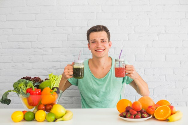 Giovane che tiene frullato verde e rosso in mano con cibo sano sul tavolo