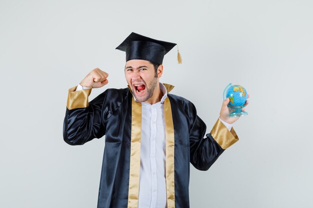 Giovane che tiene il globo della scuola in uniforme laureato e che sembra beato, vista frontale.