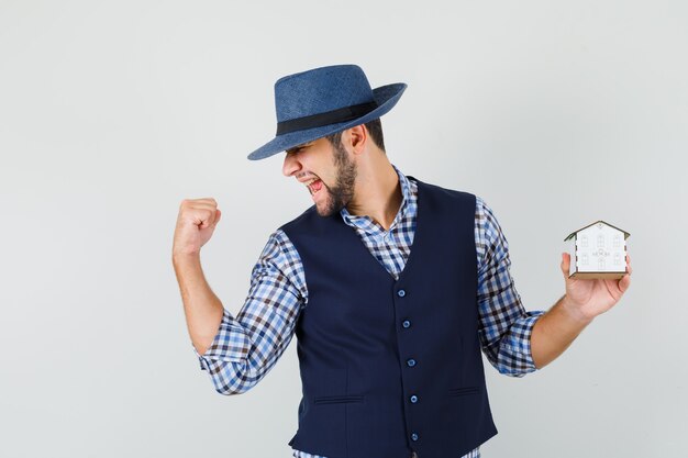 Giovane che tiene il modello della casa, mostrando il gesto del vincitore in camicia, gilet, cappello e sembra felice. vista frontale.