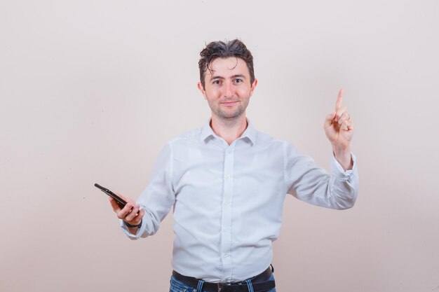 Giovane che tiene il telefono cellulare, che punta in alto in camicia, jeans e sembra positivo