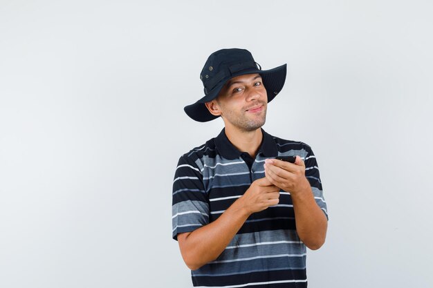 Giovane che tiene il telefono cellulare in maglietta, cappello e sembra allegro, vista frontale.