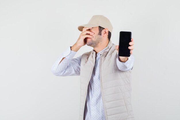 Giovane che tiene il telefono in una mano e copre parte del viso con una mano in giacca e berretto beige e sembra infastidito. vista frontale.