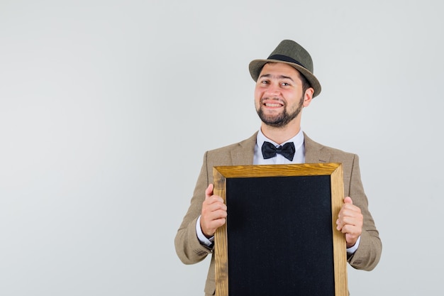 Giovane che tiene lavagna vuota in vestito, cappello e che sembra allegro, vista frontale.