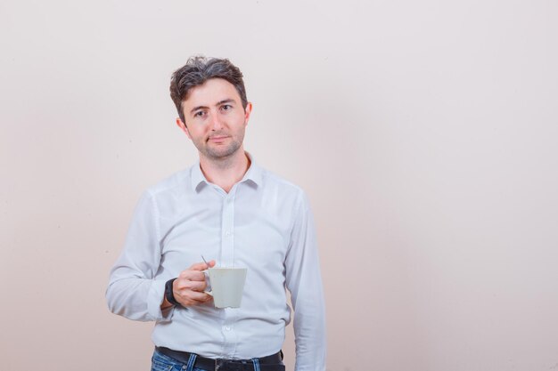 Giovane che tiene tazza di bevanda in camicia bianca, jeans e sembra pensieroso