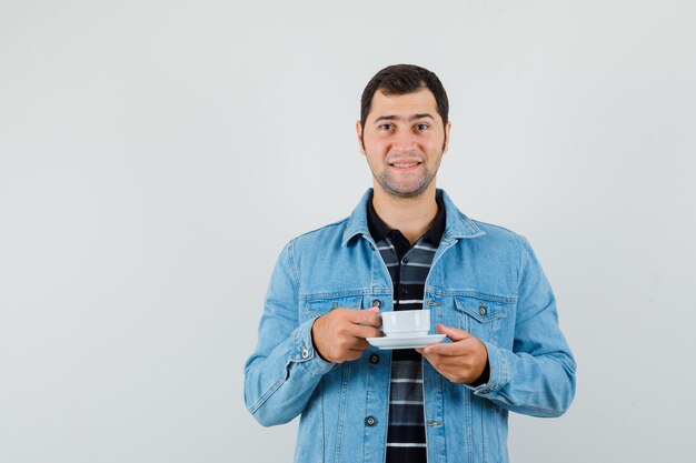 Giovane che tiene tazza di tè in maglietta, giacca e sembra allegro.