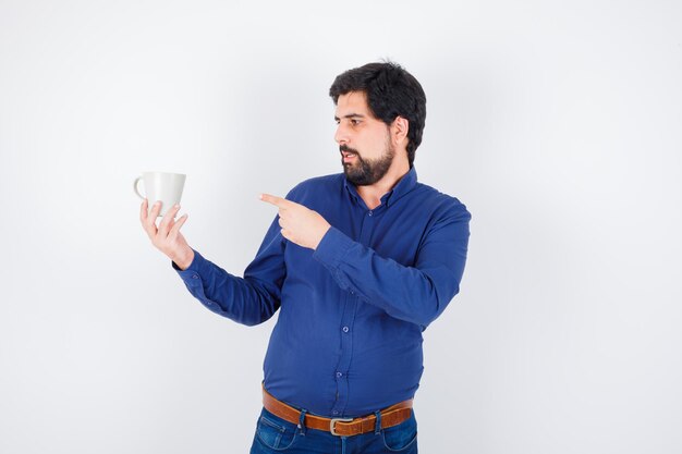 Giovane che tiene tazza e indicandolo in camicia blu e jeans e sembra ottimista. vista frontale.