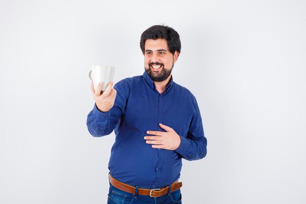 Giovane che tiene tazza e lo guarda in camicia blu e jeans e sembra ottimista, vista frontale.