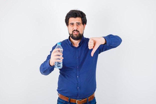 Giovane che tiene una bottiglia d'acqua e mostra il pollice giù in camicia blu e jeans e sembra serio. vista frontale.