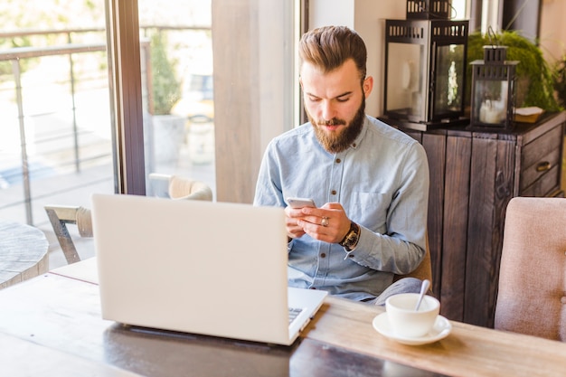 Giovane che utilizza telefono cellulare con il computer portatile e tazza di caffè sulla scrivania