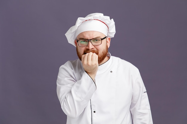 Giovane chef accigliato che indossa occhiali uniformi e berretto guardando la fotocamera mentre morde le dita isolate su sfondo viola