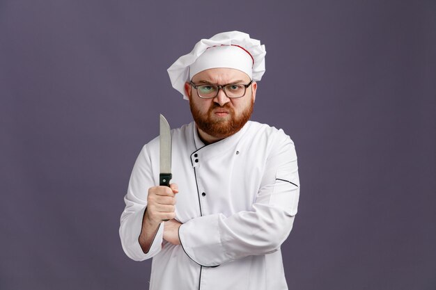 Giovane chef aggressivo che indossa occhiali uniformi e cappuccio che tiene il coltello guardando la fotocamera tenendo la mano sotto il braccio isolata su sfondo viola