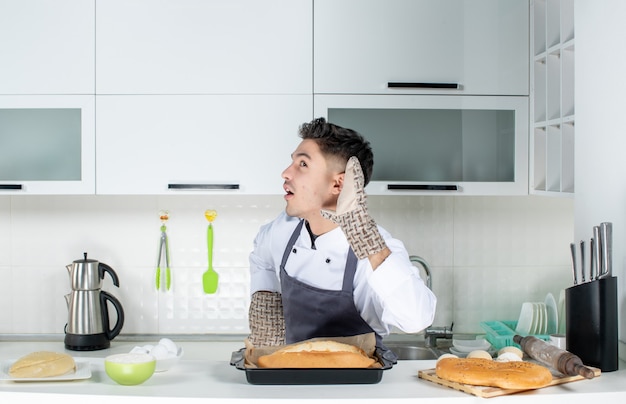 Giovane chef commis sorpreso in uniforme con supporto e pane appena sfornato sul tavolo