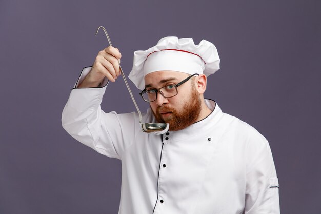 Giovane chef concentrato che indossa occhiali uniformi e cappuccio guardando la fotocamera tenendo il mestolo vicino alla bocca isolata su sfondo viola