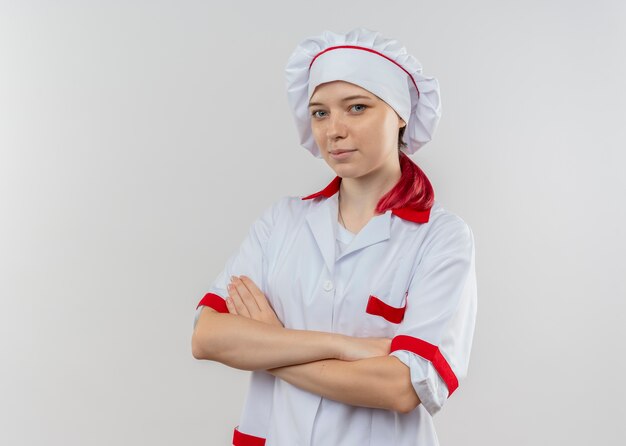 Giovane chef femmina bionda felice in uniforme del cuoco unico attraversa le braccia isolate sulla parete bianca