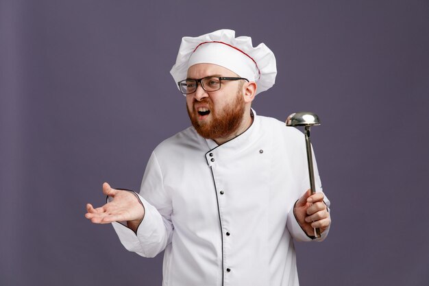 Giovane chef infastidito che indossa occhiali uniformi e cappuccio che tiene un mestolo guardando il lato che mostra la mano vuota isolata su sfondo viola