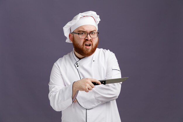 Giovane chef infastidito che indossa occhiali uniformi e cappuccio guardando la fotocamera che punta di lato con il coltello isolato su sfondo viola