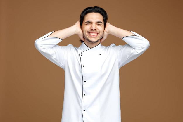 giovane chef irritato che indossa le orecchie di chiusura uniformi con le mani con gli occhi chiusi isolati su sfondo marrone