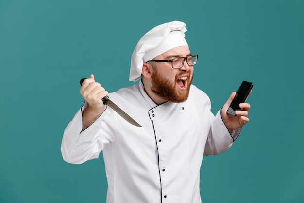 Giovane chef maschio arrabbiato che indossa l'uniforme degli occhiali e il cappuccio che tiene coltello e telefono cellulare guardando il lato gridando isolato su sfondo blu