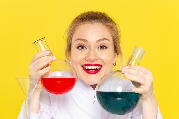 Giovane chimico femminile di vista frontale in vestito bianco che tiene soluzioni differenti che sorridono sul lavoro di chimica dello spazio giallo