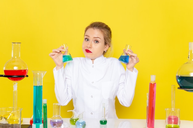 Giovane chimico femminile di vista frontale in vestito bianco davanti al tavolo con soluzioni ed lavorando con loro sul lavoro di chimica dello spazio giallo