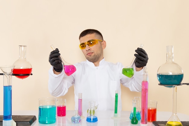 Giovane chimico maschio di vista frontale in vestito speciale bianco che si siede davanti al tavolo con soluzioni che tengono sull'esperimento di scienza di chimica del laboratorio della parete crema
