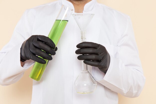 Giovane chimico maschio di vista ravvicinata anteriore in vestito speciale bianco che tiene piccola boccetta con soluzione verde sull'esperimento di scienza di chimica del laboratorio dello scrittorio crema