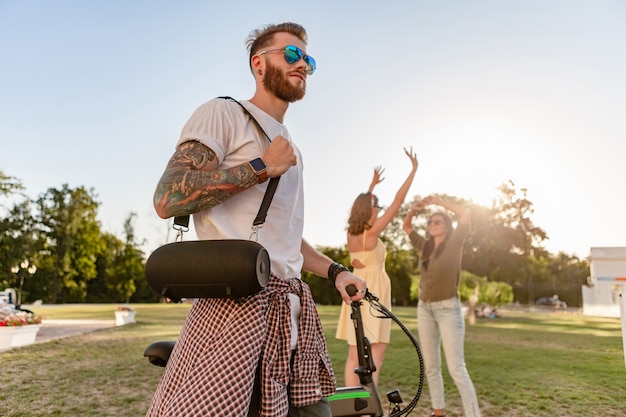 Giovane compagnia hipster di amici che si divertono insieme nel parco sorridendo ascoltando musica su altoparlanti wireless