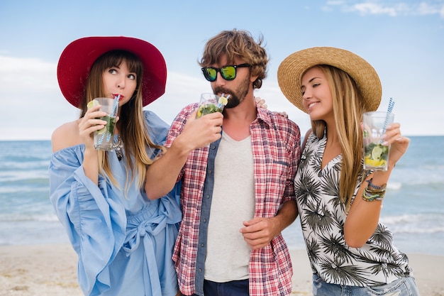 Giovane compagnia hipster di amici in vacanza in spiaggia, bevendo cocktail mojito, felice positivo, stile estivo, sorridendo felice, due donne e un uomo che si divertono insieme, parlando, flirt, romanticismo, tre